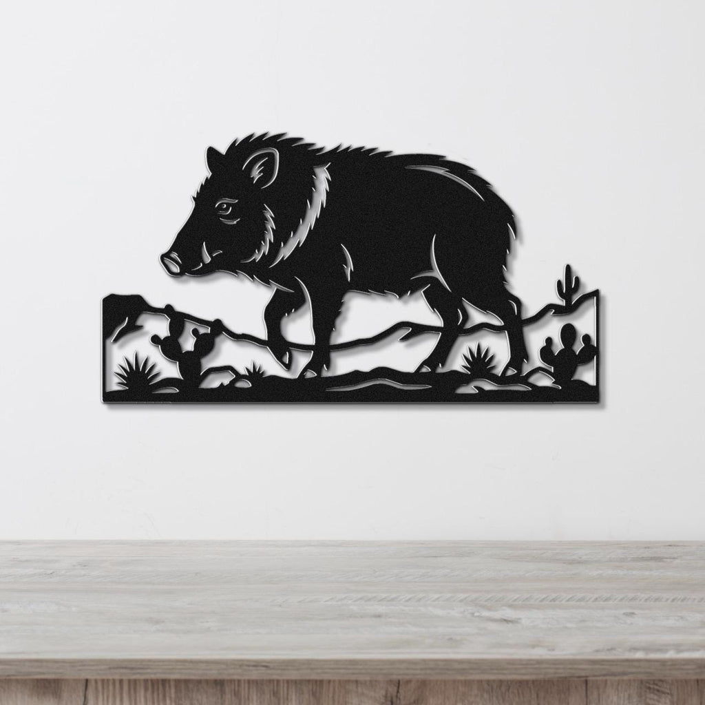 Black metal wall art featuring a javelina silhouette with cacti and desert terrain cutouts, mounted on a white wall above a wooden surface.