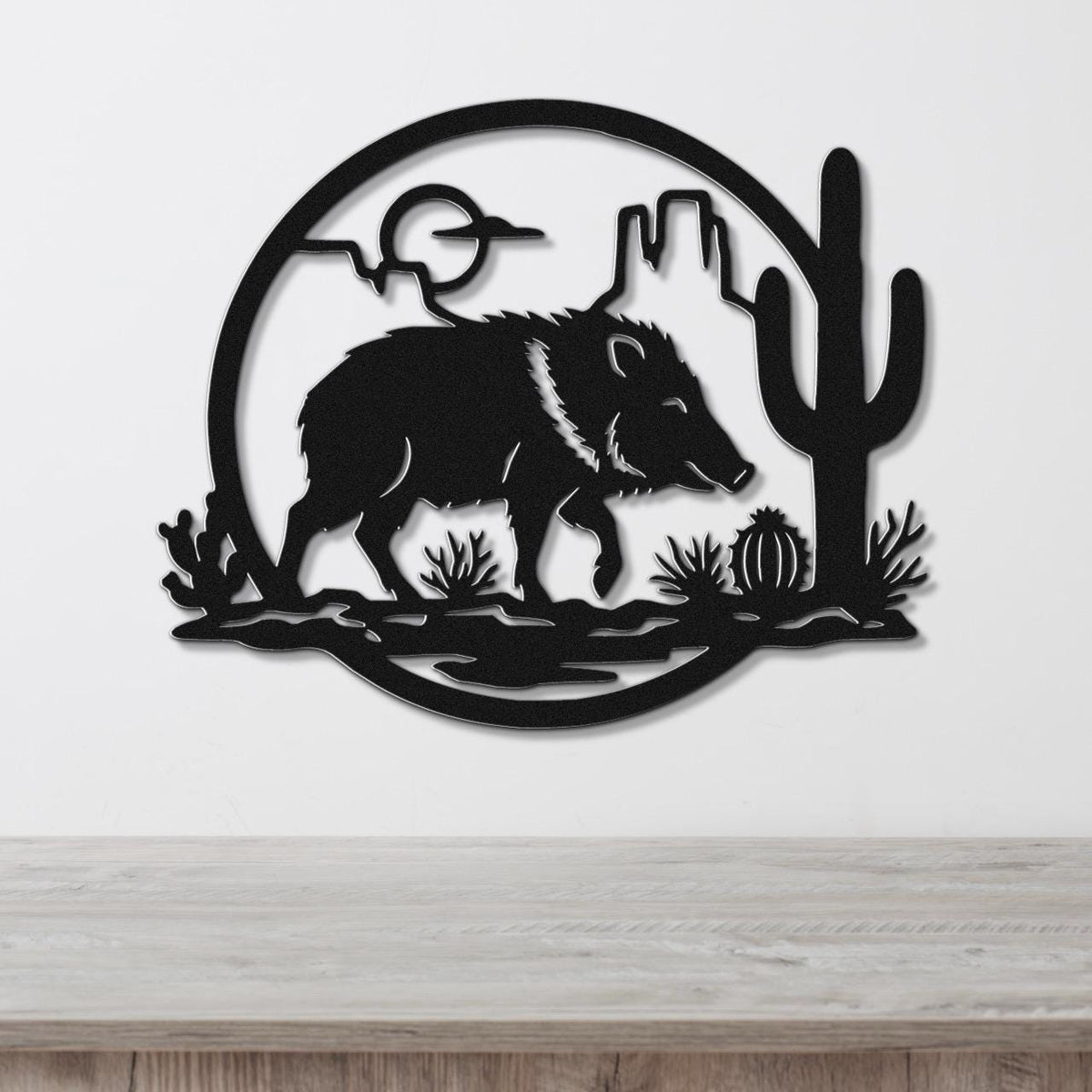Black metal wall art silhouette of a javelina in a desert scene with cactus and mountains, mounted on a white wall above a wooden surface.