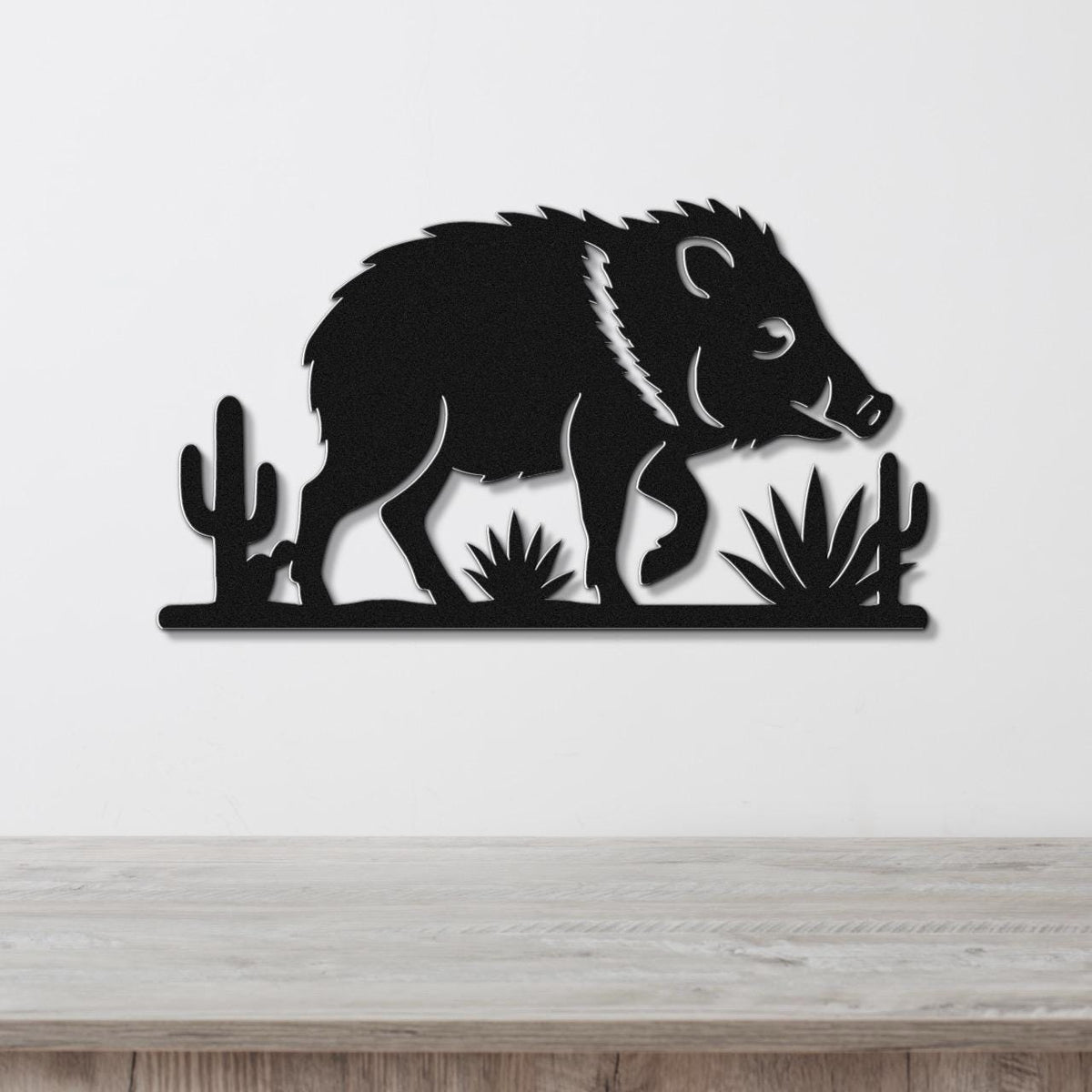 Black metal wall art cutout of a javelina with cacti and agave plants, mounted on a white wall above a wooden surface.