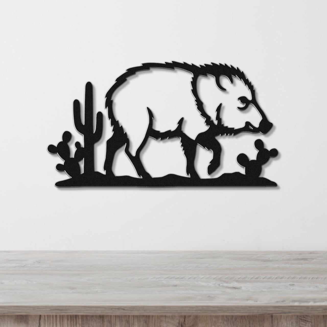 Black metal wall art silhouette of a javelina with cacti, mounted on a white wall above a wooden surface.