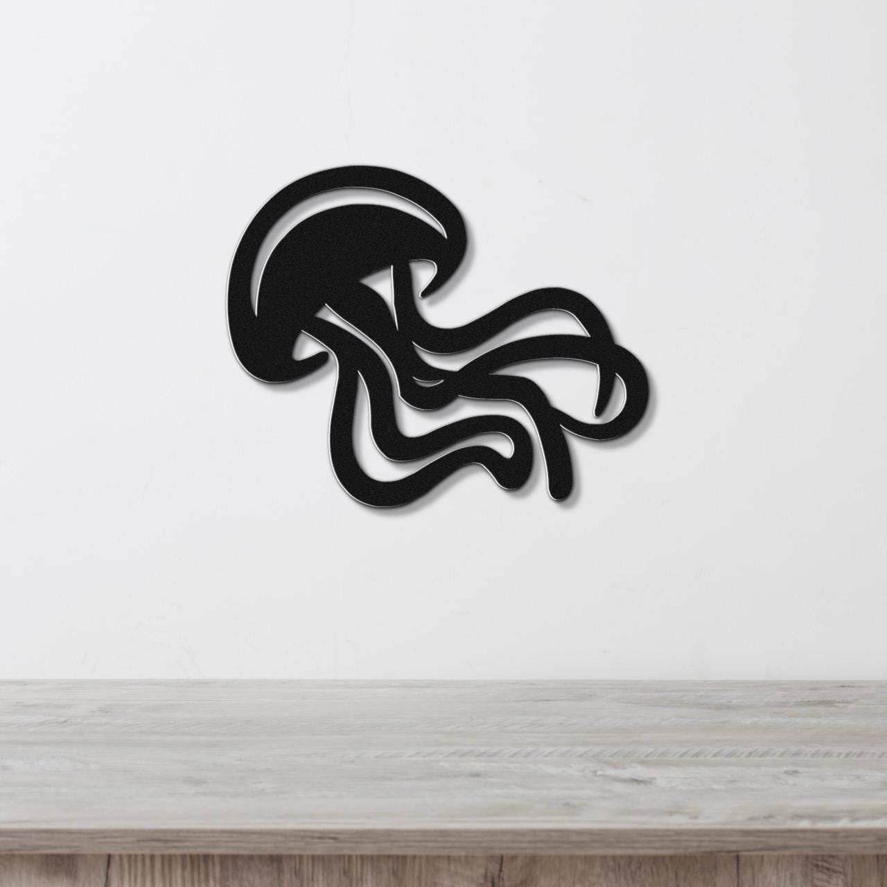 Black metal jellyfish wall art with flowing tentacle cutouts, mounted on a white wall above a light wood surface.