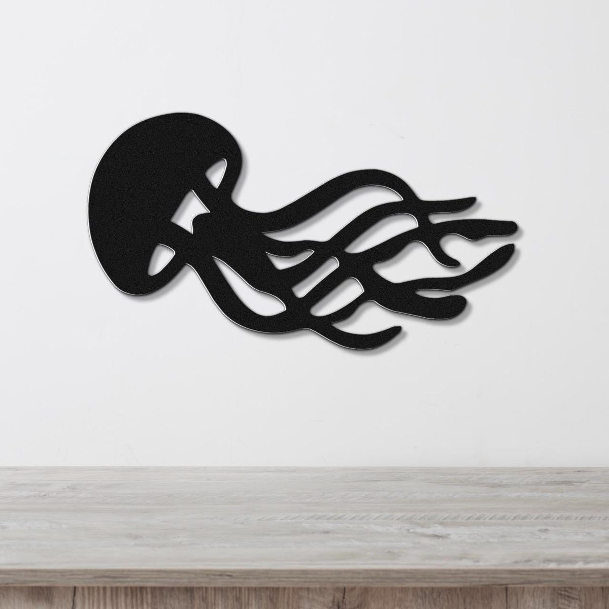 Black metal jellyfish wall art with flowing tentacle cutouts, mounted on a white wall above a light wood surface.