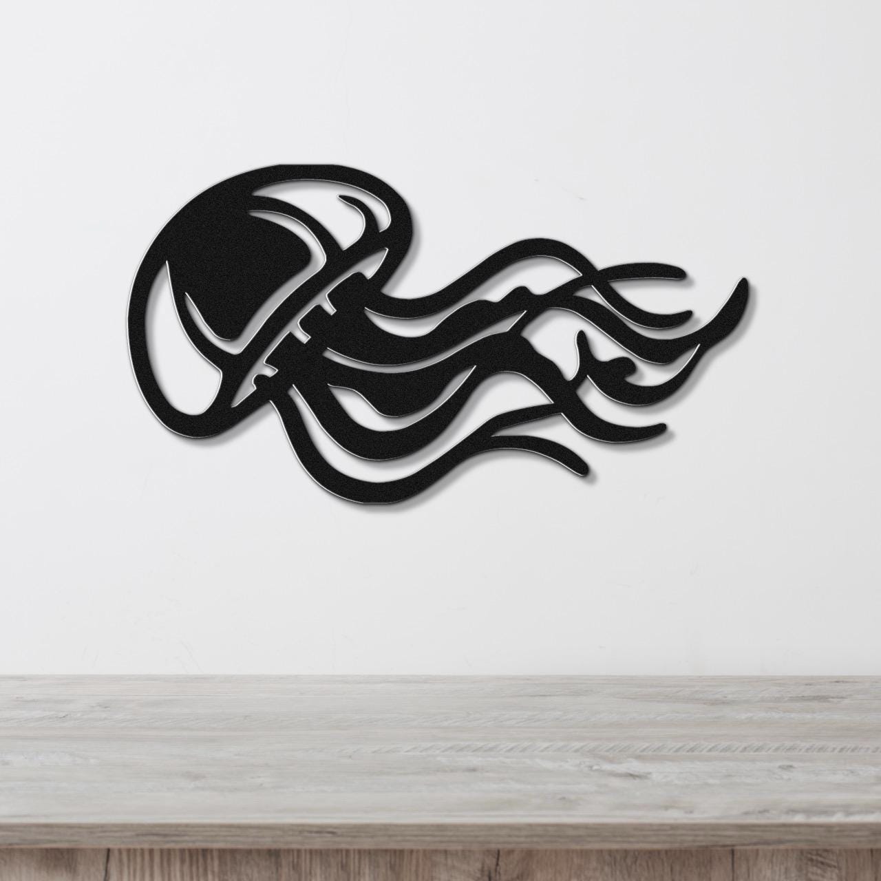 Black metal jellyfish wall art with flowing tentacle cutouts, mounted on a white wall above a light wood surface.