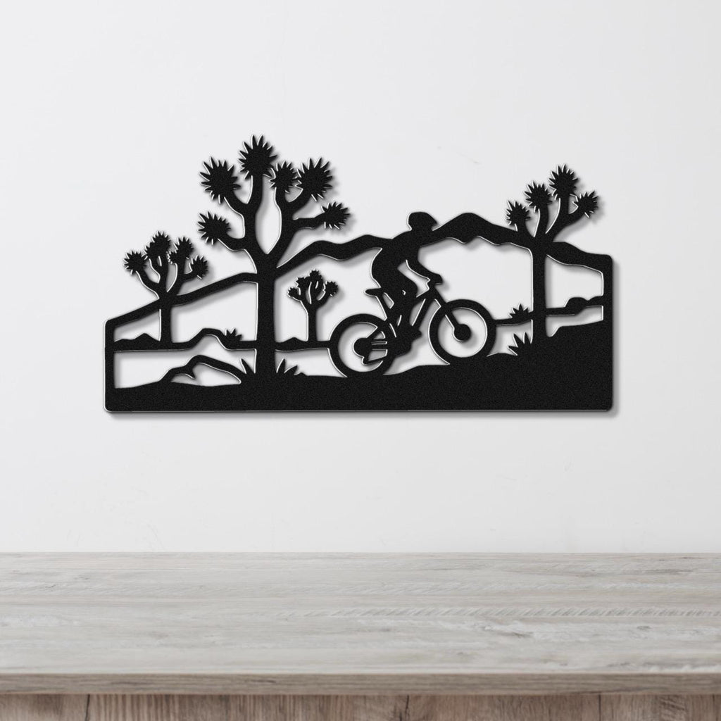 Black metal wall art featuring a cyclist silhouette and Joshua trees in a southwestern landscape, mounted on a white wall above a wooden surface.