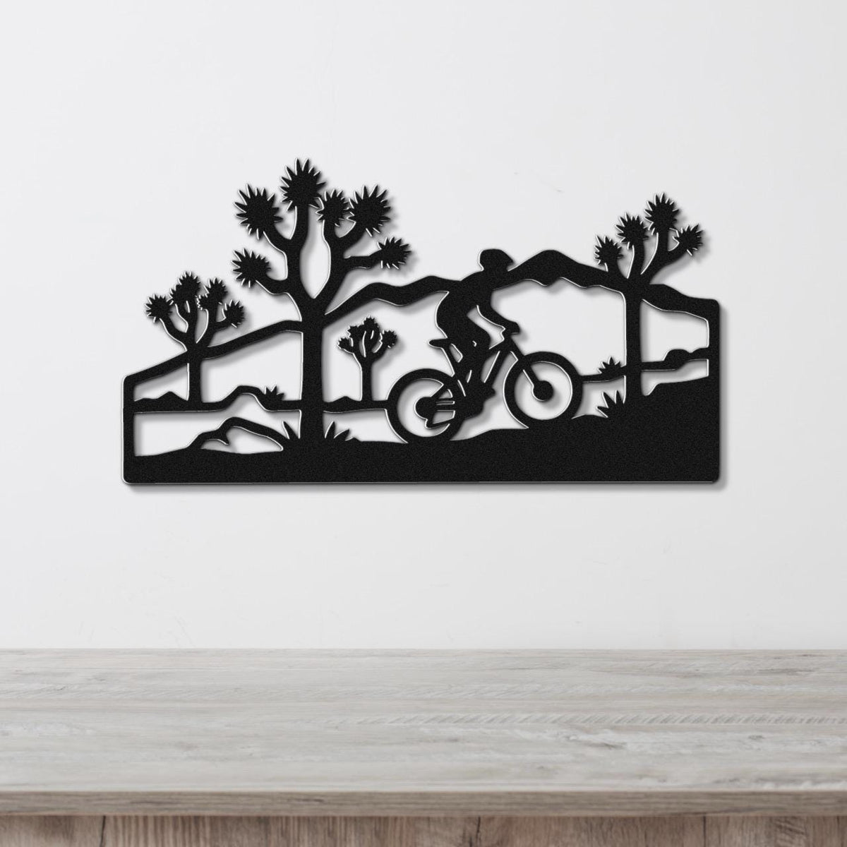 Black metal wall art featuring a cyclist silhouette and Joshua trees in a southwestern landscape, mounted on a white wall above a wooden surface.
