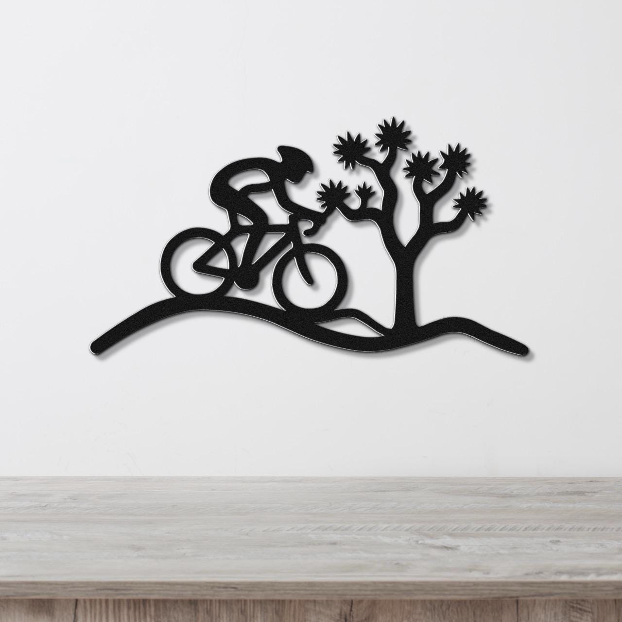 Black metal wall art silhouette of a cyclist on a desert trail with a Joshua tree, mounted on a white wall above a wooden surface.