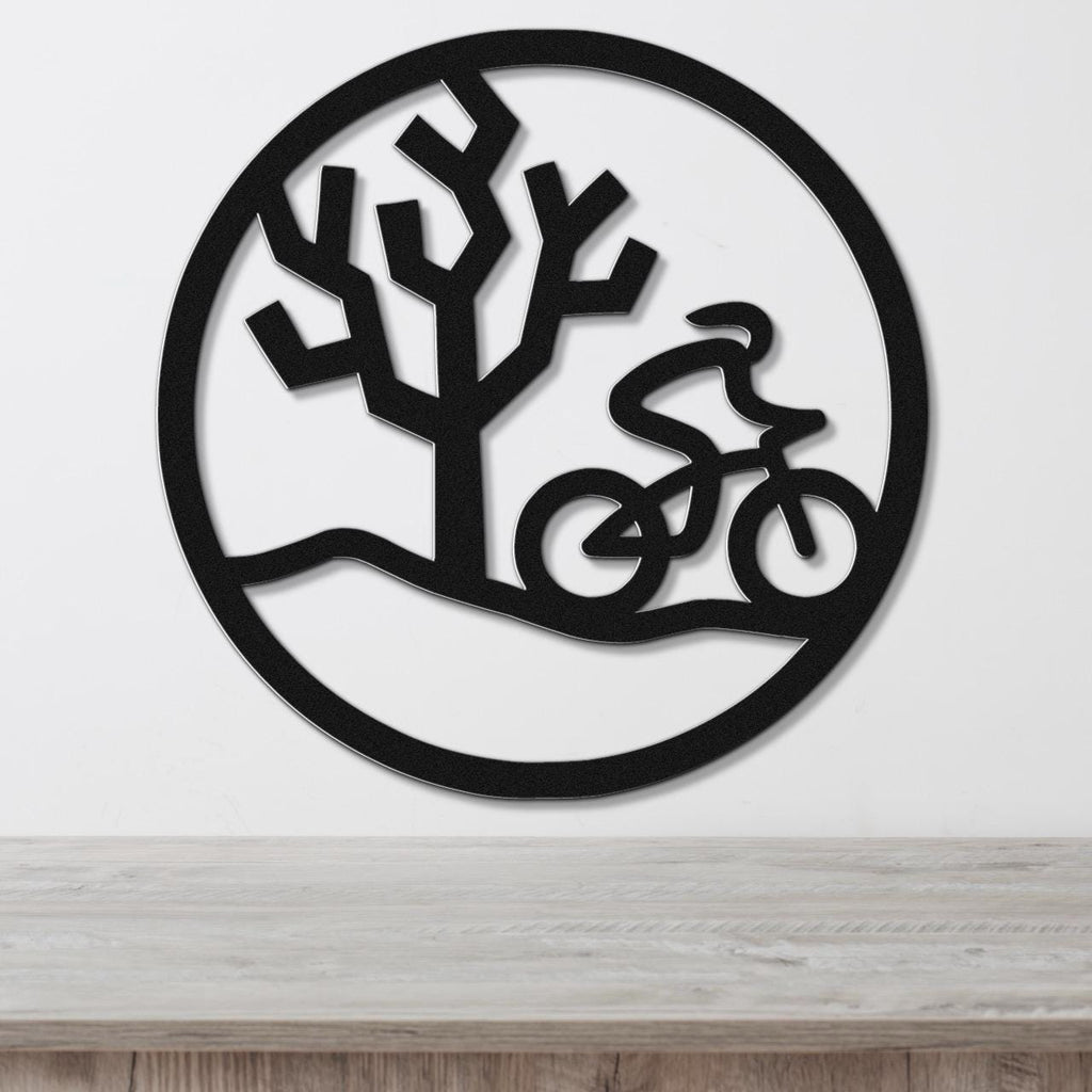 Black metal circular wall art with a cyclist in motion and a tree silhouette, mounted on a white wall above a wooden surface.