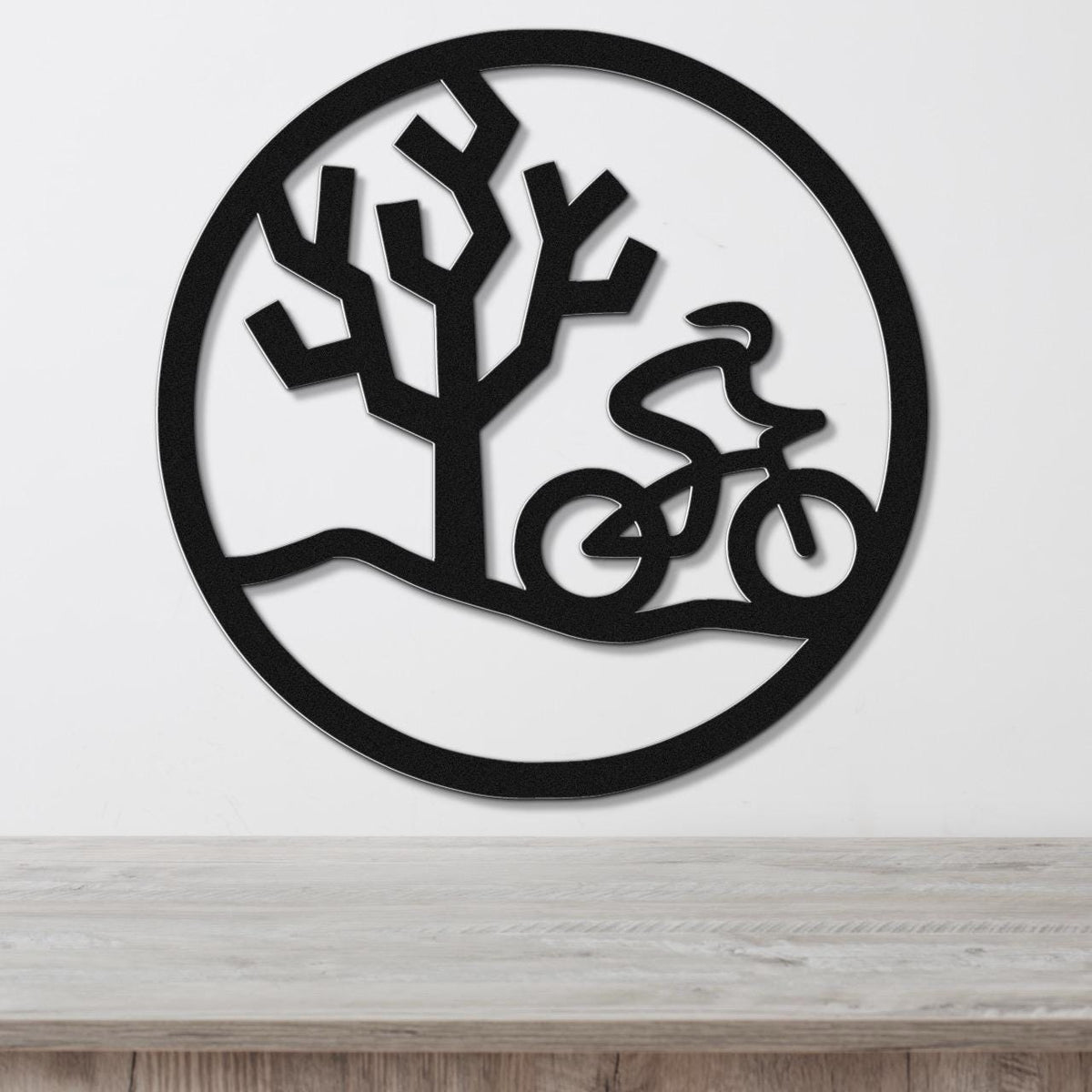 Black metal circular wall art with a cyclist in motion and a tree silhouette, mounted on a white wall above a wooden surface.