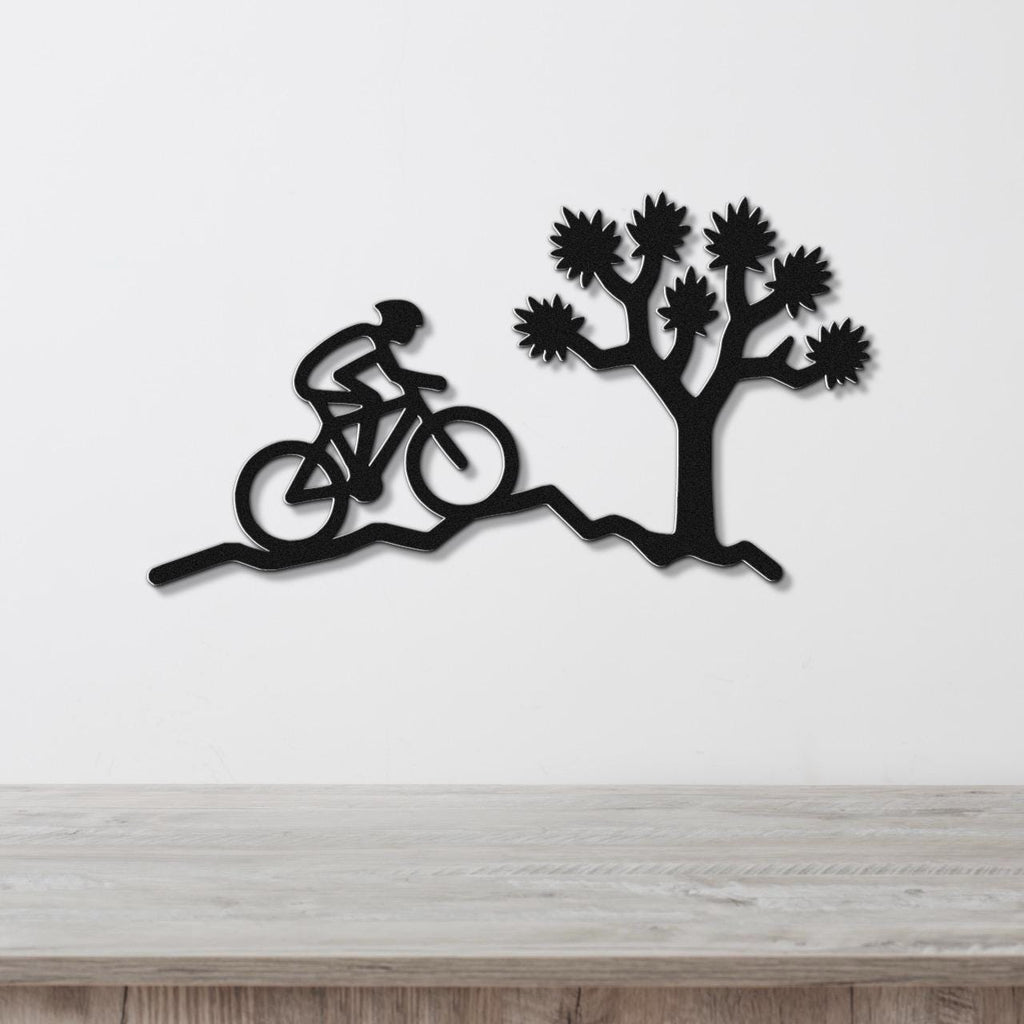 Black metal wall art cutout of a cyclist on a trail beside a Joshua tree, mounted on a white wall above a wooden surface.