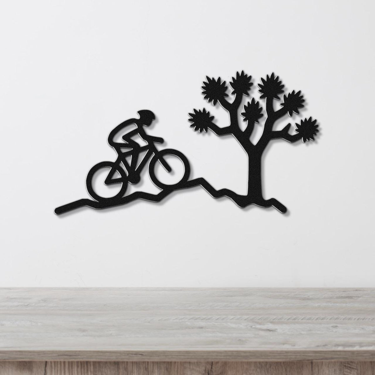 Black metal wall art cutout of a cyclist on a trail beside a Joshua tree, mounted on a white wall above a wooden surface.