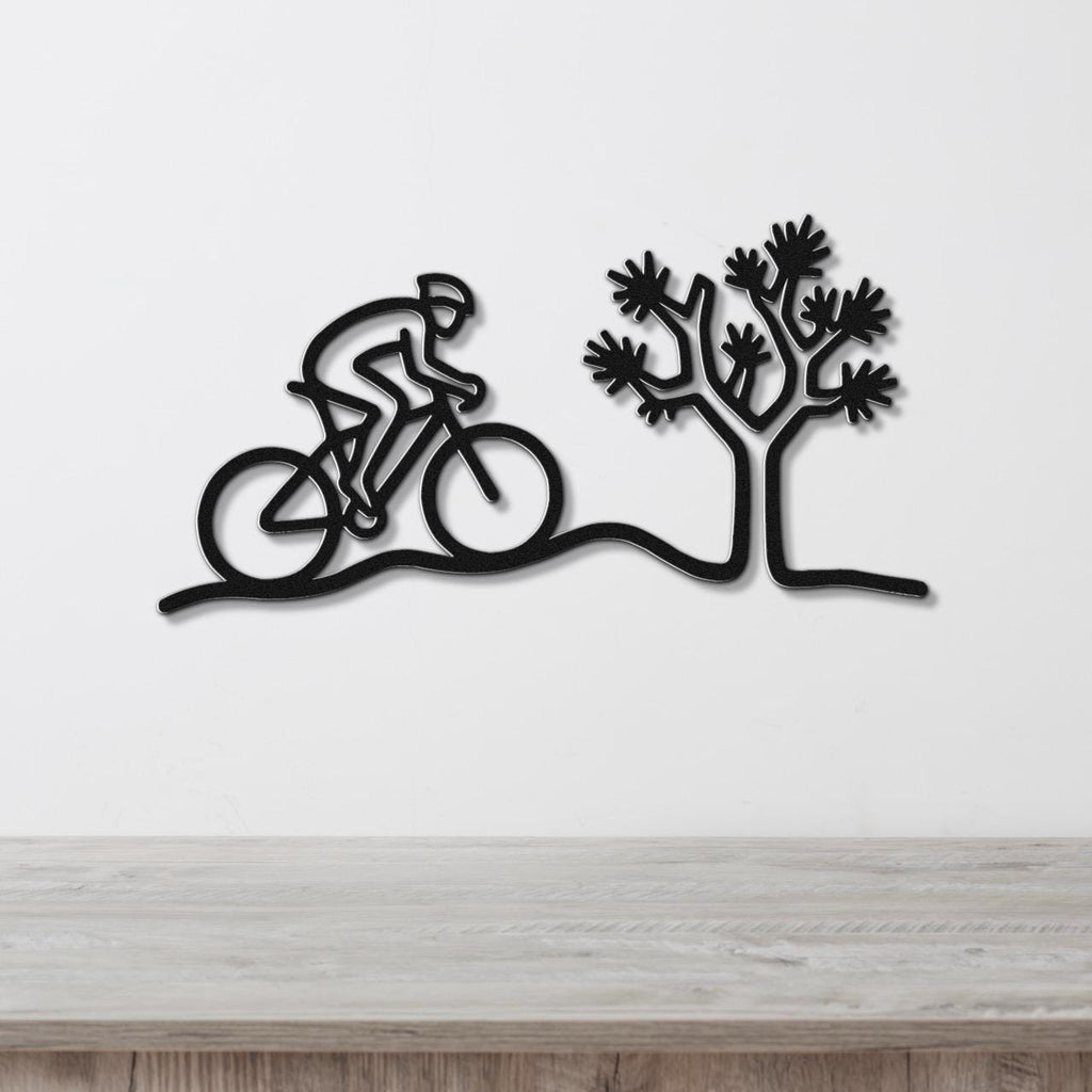 Black metal wall art silhouette of a cyclist on a desert trail with a Joshua tree, mounted on a white wall above a wooden surface.