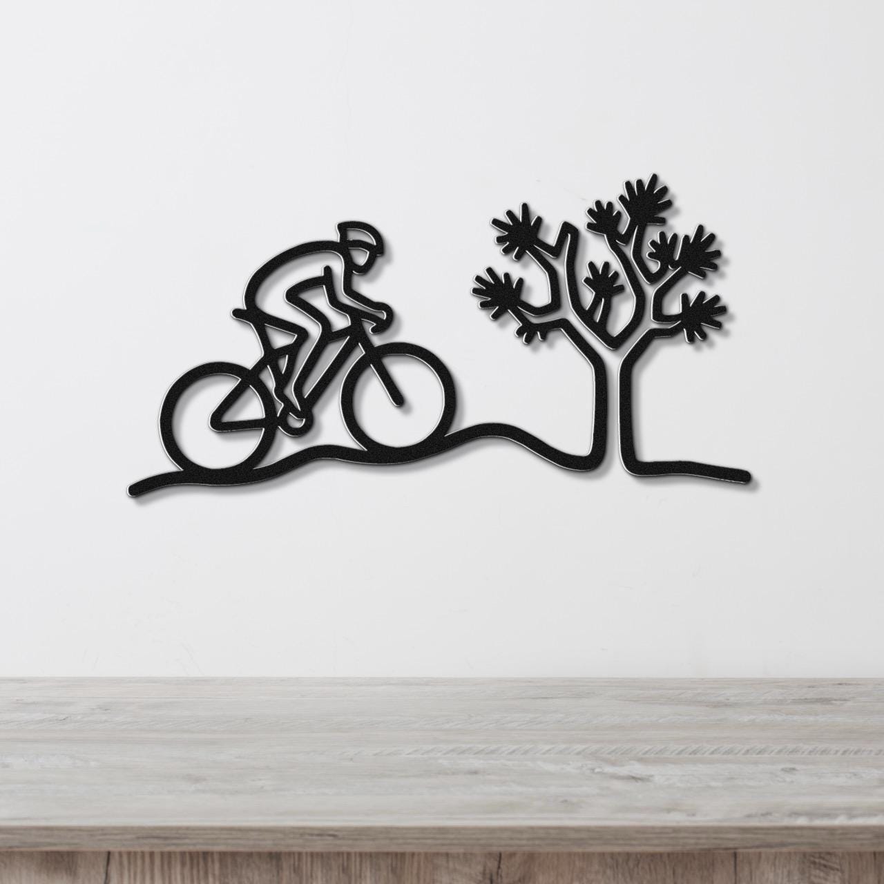 Black metal wall art silhouette of a cyclist on a desert trail with a Joshua tree, mounted on a white wall above a wooden surface.