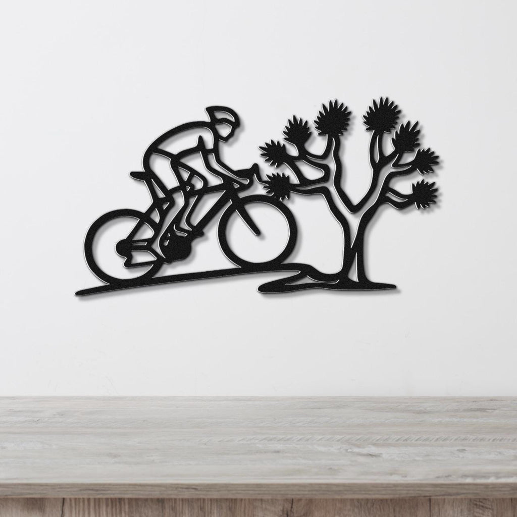 Black metal wall art cutout of a cyclist riding past a Joshua tree, mounted on a white wall above a wooden surface.