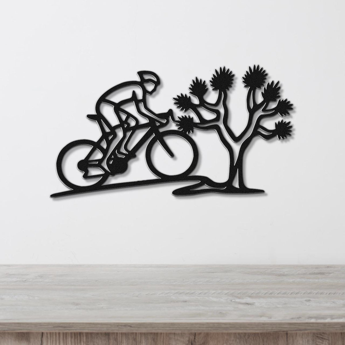 Black metal wall art cutout of a cyclist riding past a Joshua tree, mounted on a white wall above a wooden surface.
