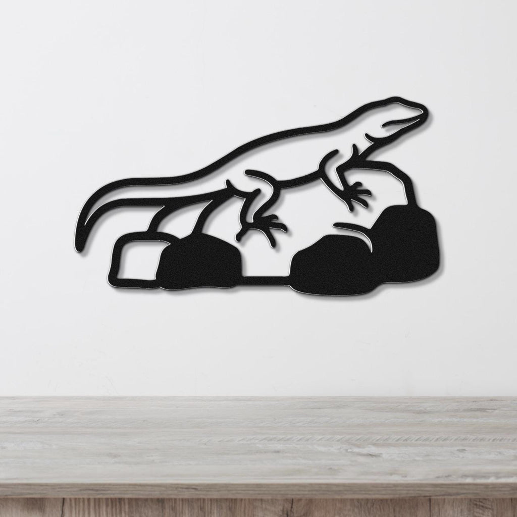 Black metal wall art featuring a lizard silhouette perched on rocks, mounted on a white wall above a wooden surface.