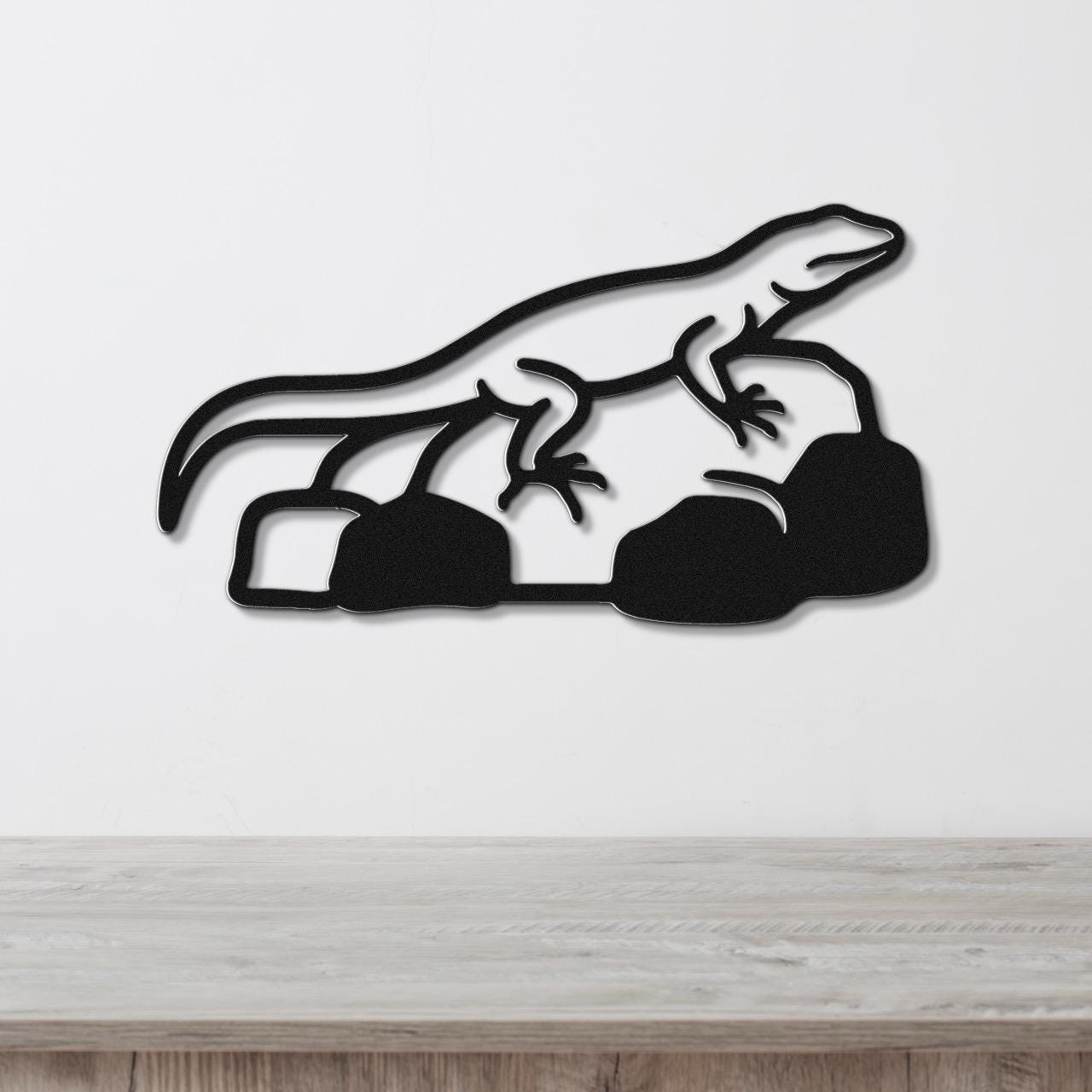 Black metal wall art featuring a lizard silhouette perched on rocks, mounted on a white wall above a wooden surface.