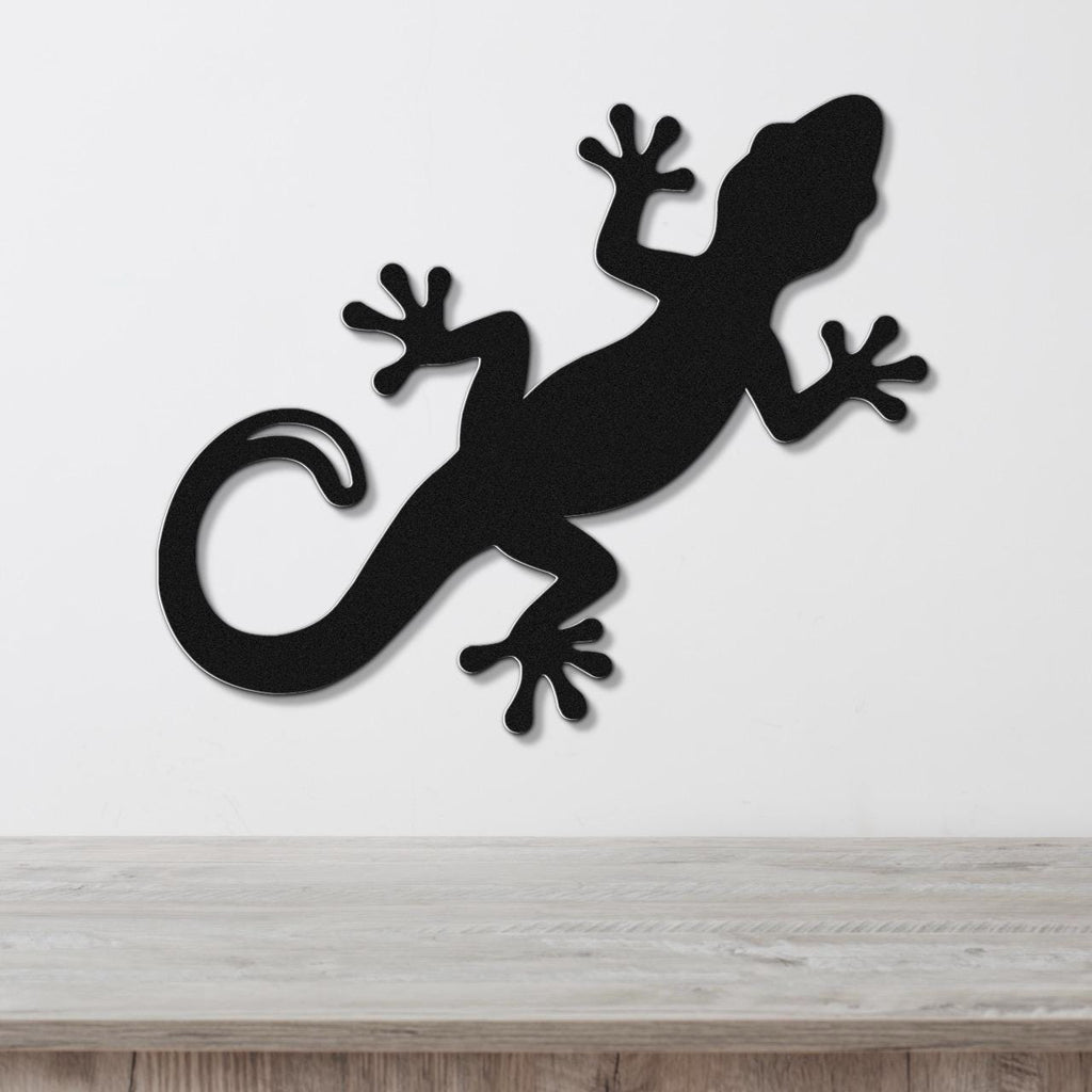 Black metal gecko wall art with a silhouette cutout design, mounted on a white wall above a light wood surface.