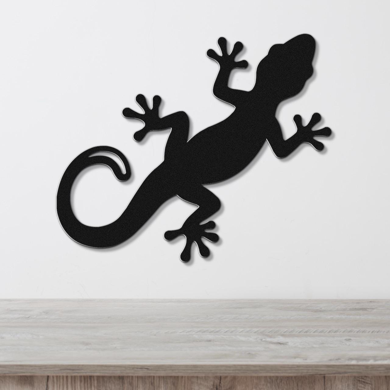 Black metal gecko wall art with a silhouette cutout design, mounted on a white wall above a light wood surface.