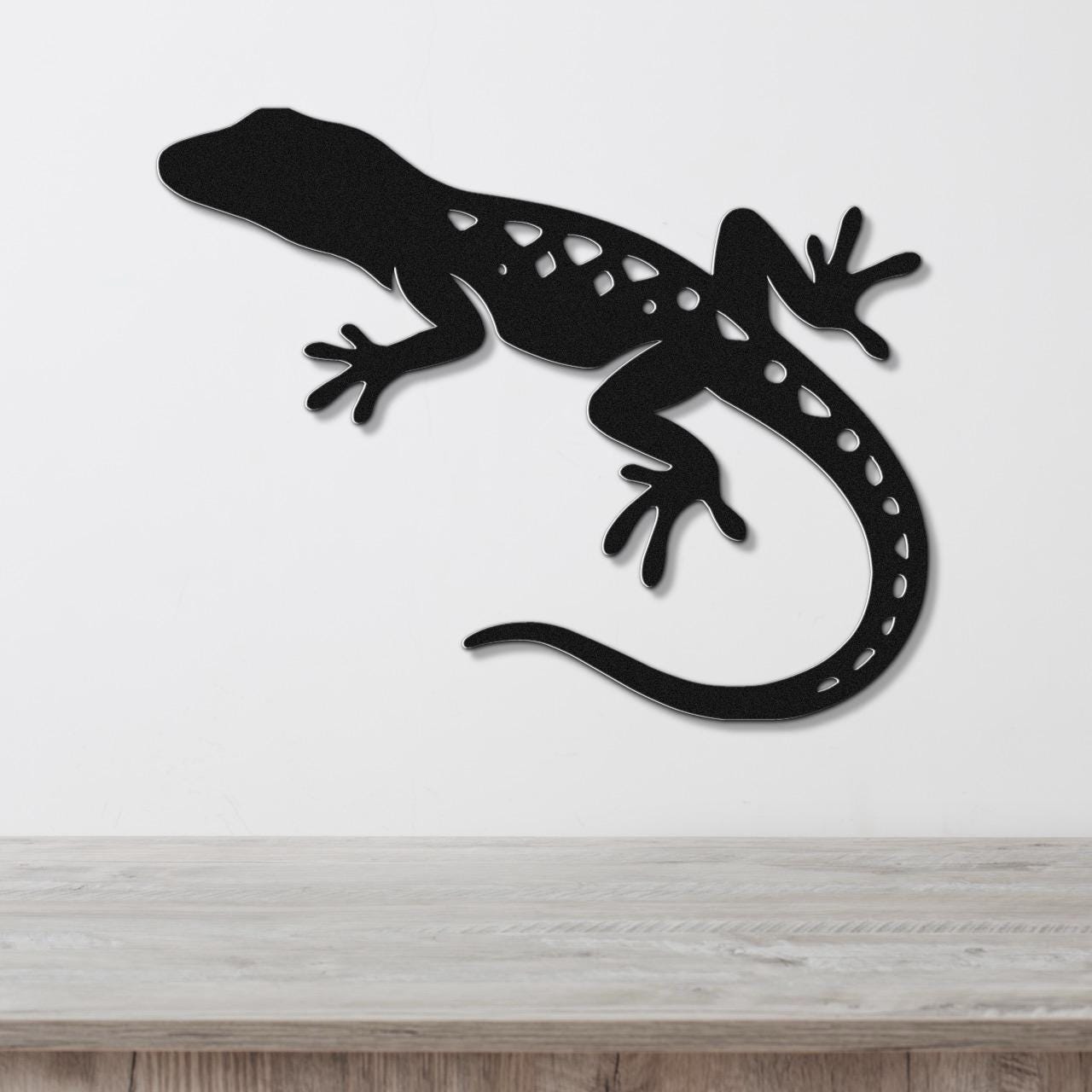 Black metal lizard wall art with cutout details, mounted on a white wall above a wooden surface.