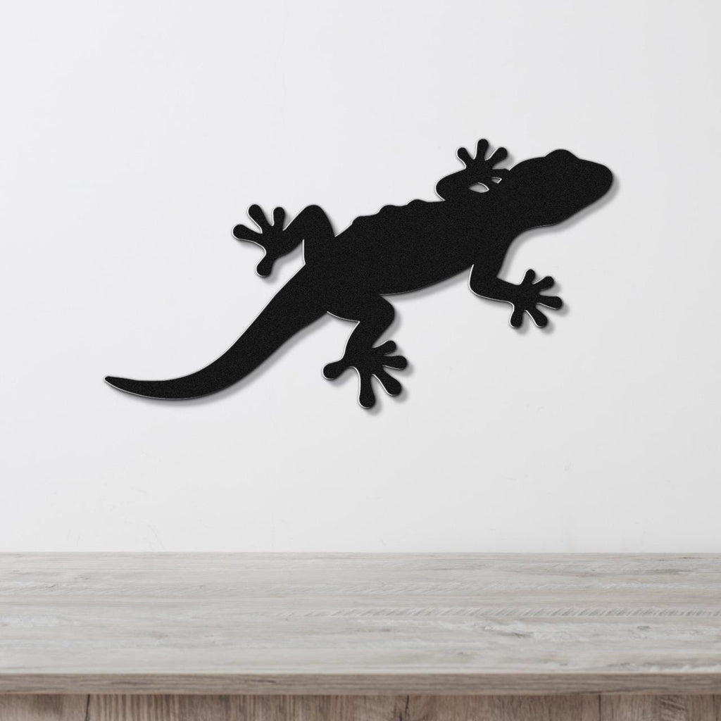 Black metal lizard wall art silhouette with detailed cutout feet and tail, mounted on a white wall above a light wood surface.