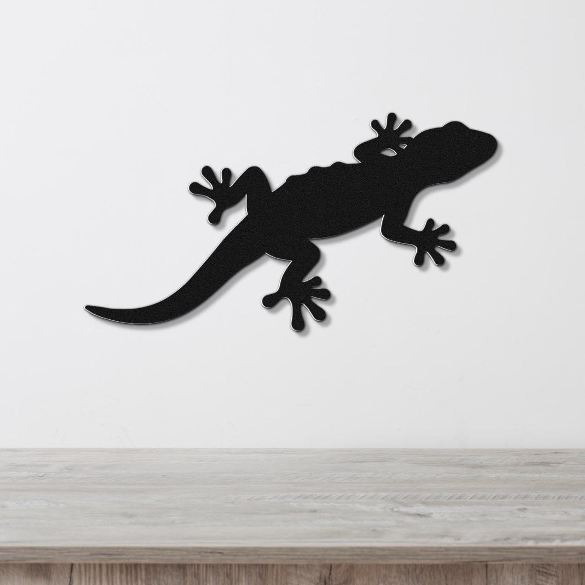 Black metal lizard wall art silhouette with detailed cutout feet and tail, mounted on a white wall above a light wood surface.