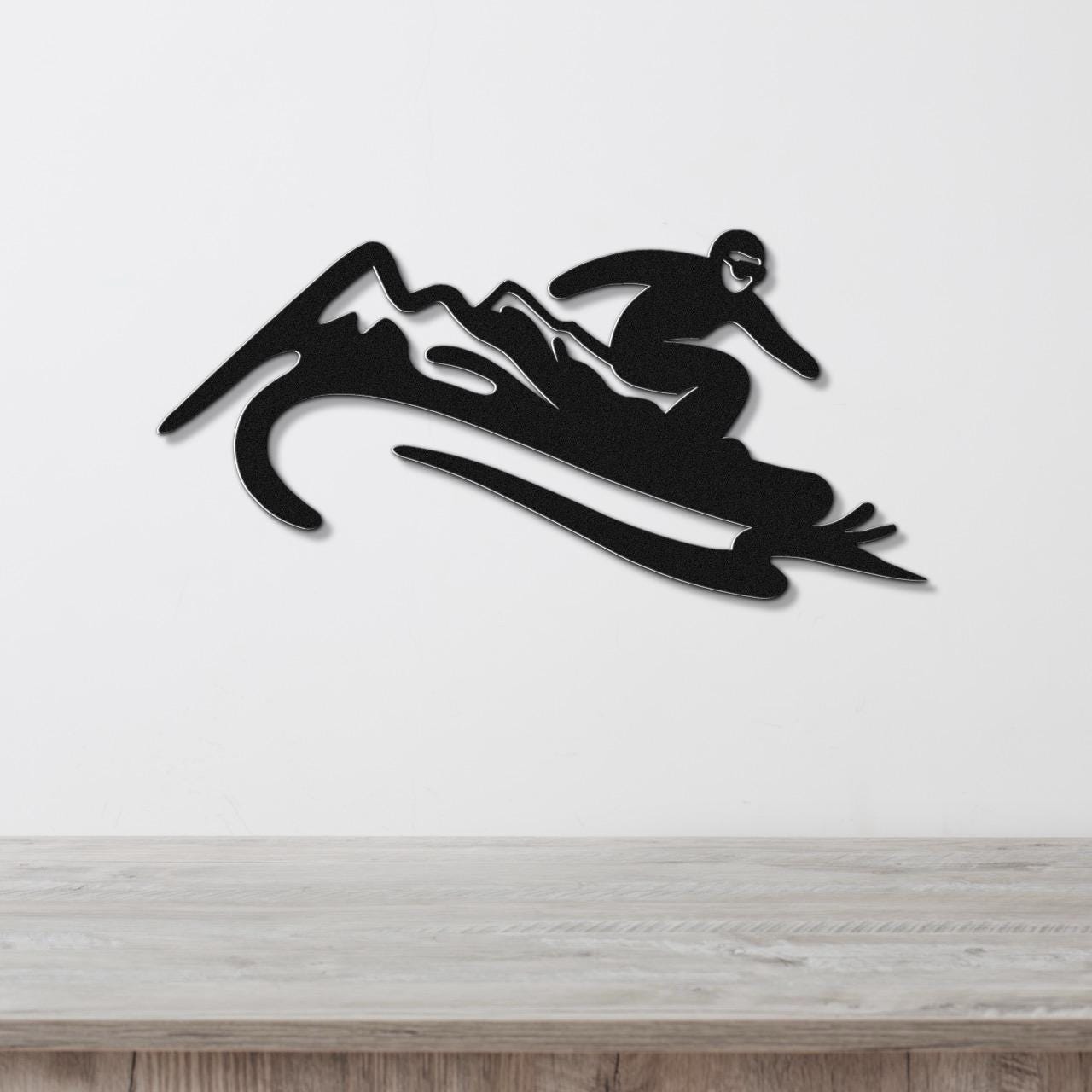 Black metal wall art cutout of a snowboarder in motion over mountain peaks, mounted on a white wall above a wooden surface.