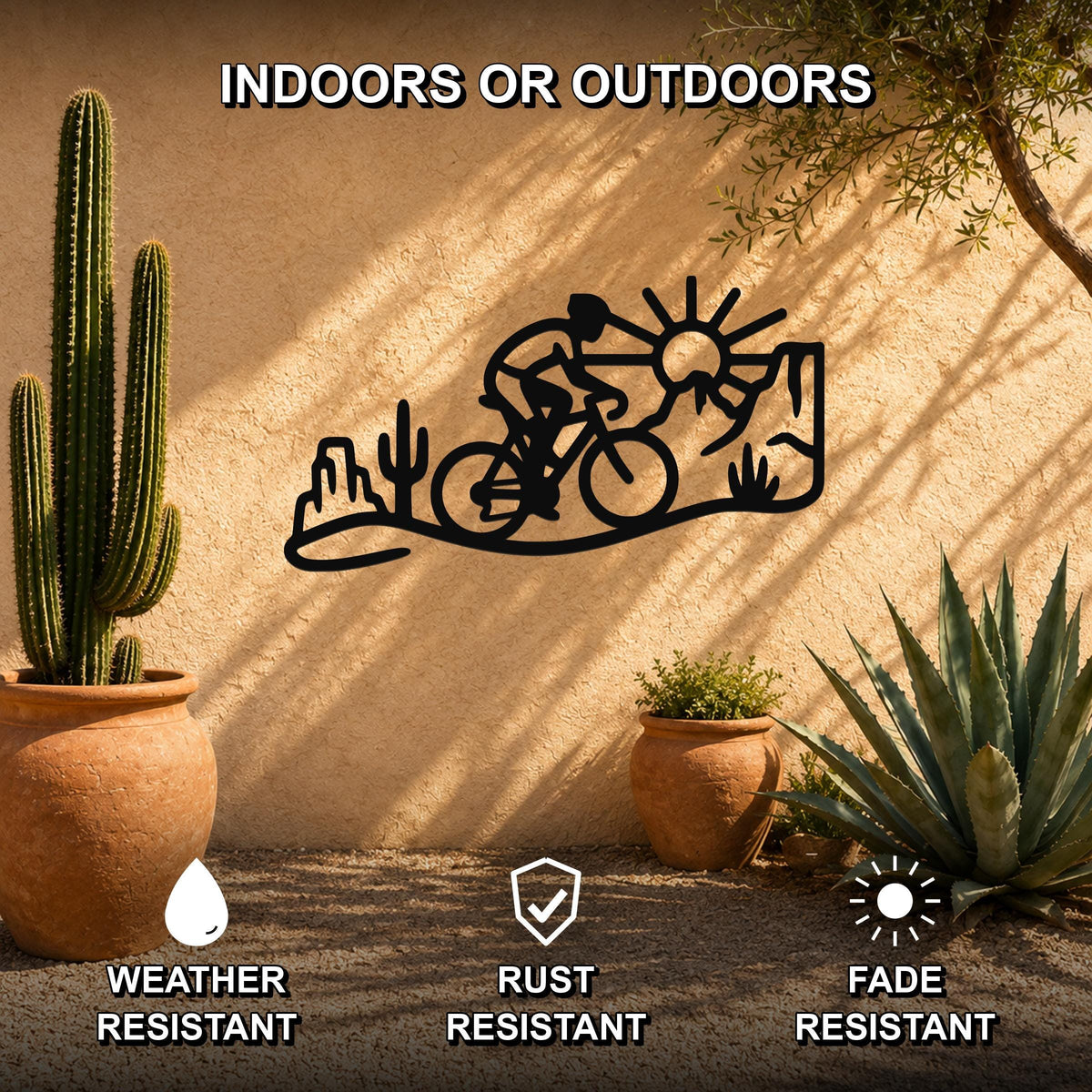 Black metal wall art silhouette of a cyclist in a southwestern desert scene with cacti and sun, mounted on an outdoor stucco wall between potted plants.