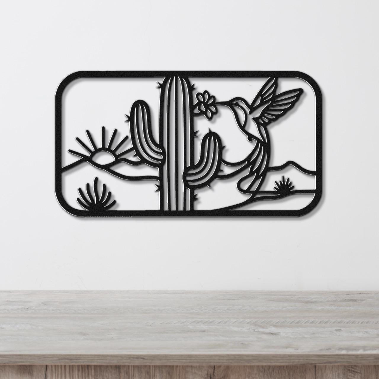 Black metal wall art with a cutout design of a saguaro cactus, hummingbird, and desert flowers, mounted on a white wall above a wooden surface.