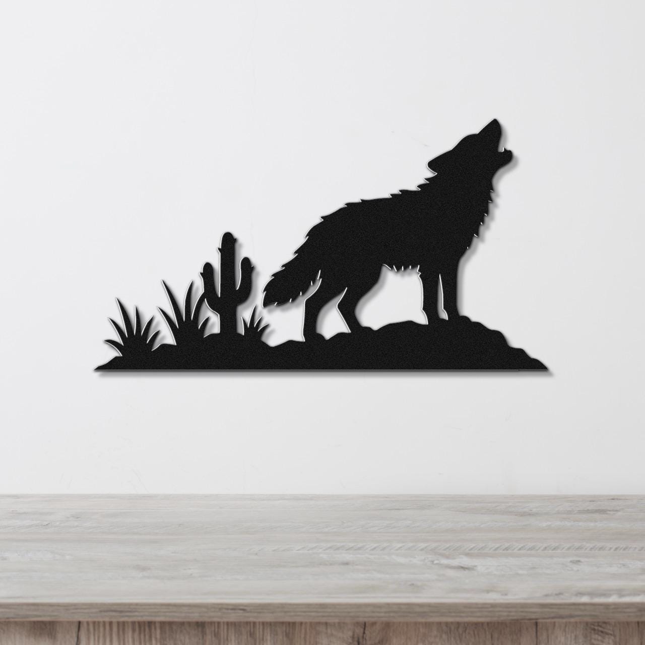 Black metal wall art silhouette of a coyote howling on a rock with cactus and desert plants, mounted on a white wall above a wooden surface.
