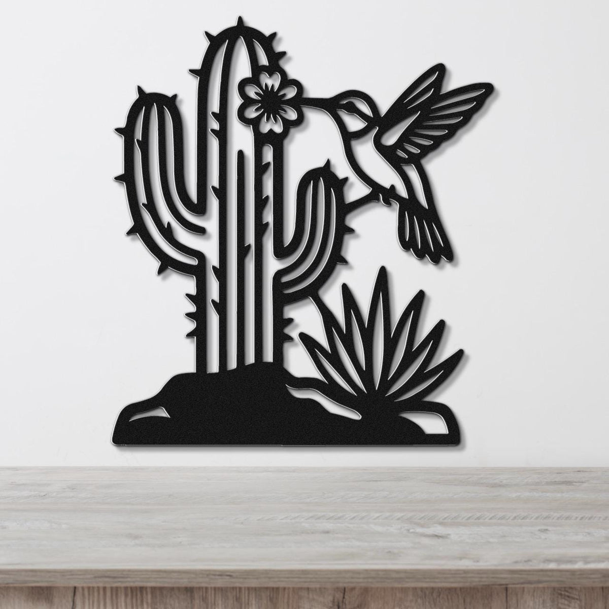 Black metal wall art featuring a cactus, hummingbird, and flower cutouts, mounted on a white wall above a wooden surface.