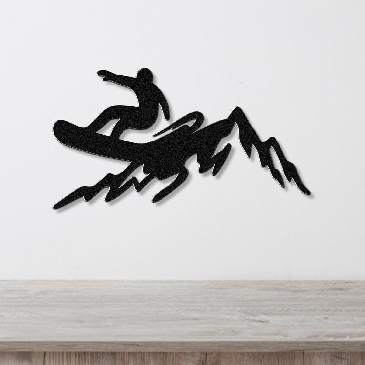 Black metal wall art silhouette of a snowboarder in mid-air above a mountain range, mounted on a white wall above a light wood counter.