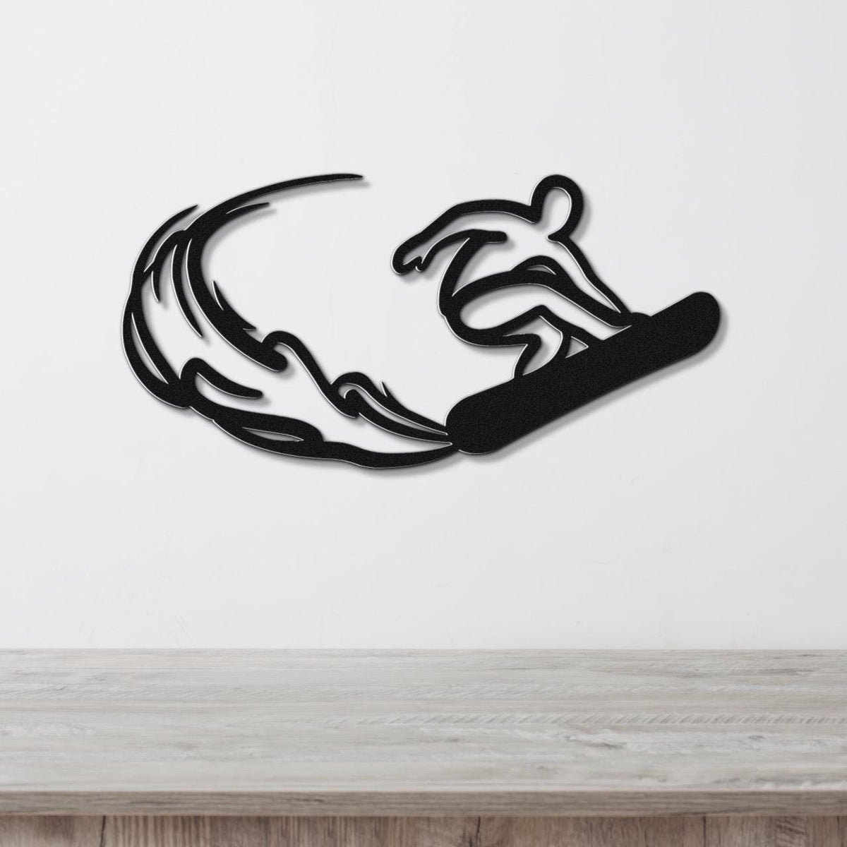 Black metal wall art silhouette of a snowboarder in motion with wave-like cutouts, mounted on a white wall above a wooden surface.