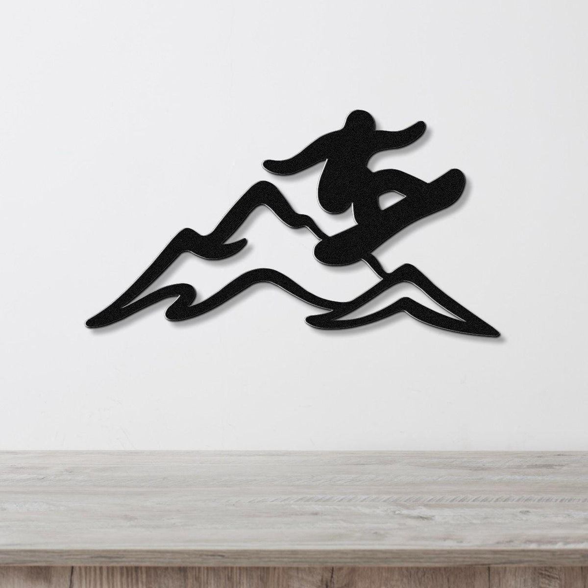 Black metal wall art silhouette of a snowboarder in motion over mountain peaks, mounted on a white wall above a wooden surface.