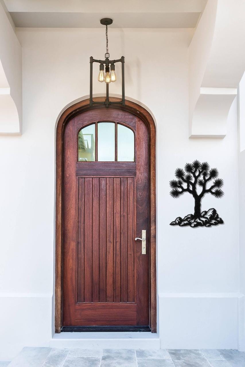 Black metal wall art of a Joshua tree silhouette with rocky base, mounted on a white wall beside a wooden front door.