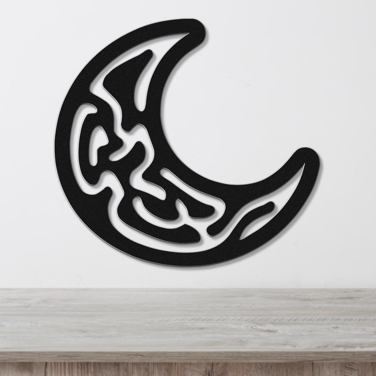 Black metal crescent moon wall art with intricate internal cutout patterns, mounted on a white wall above a light wood surface.