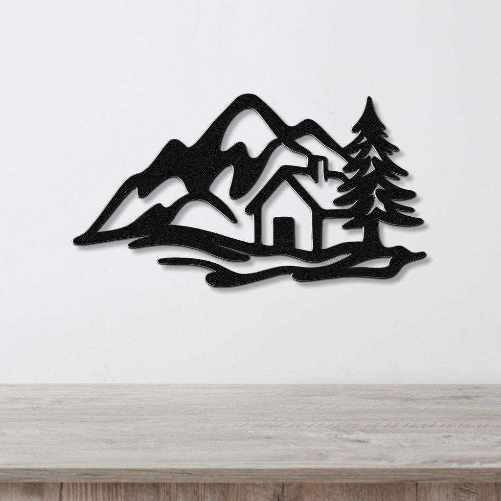 Black metal wall art silhouette of a mountain cabin scene with pine trees and peaks, mounted on a white wall above a wooden surface.