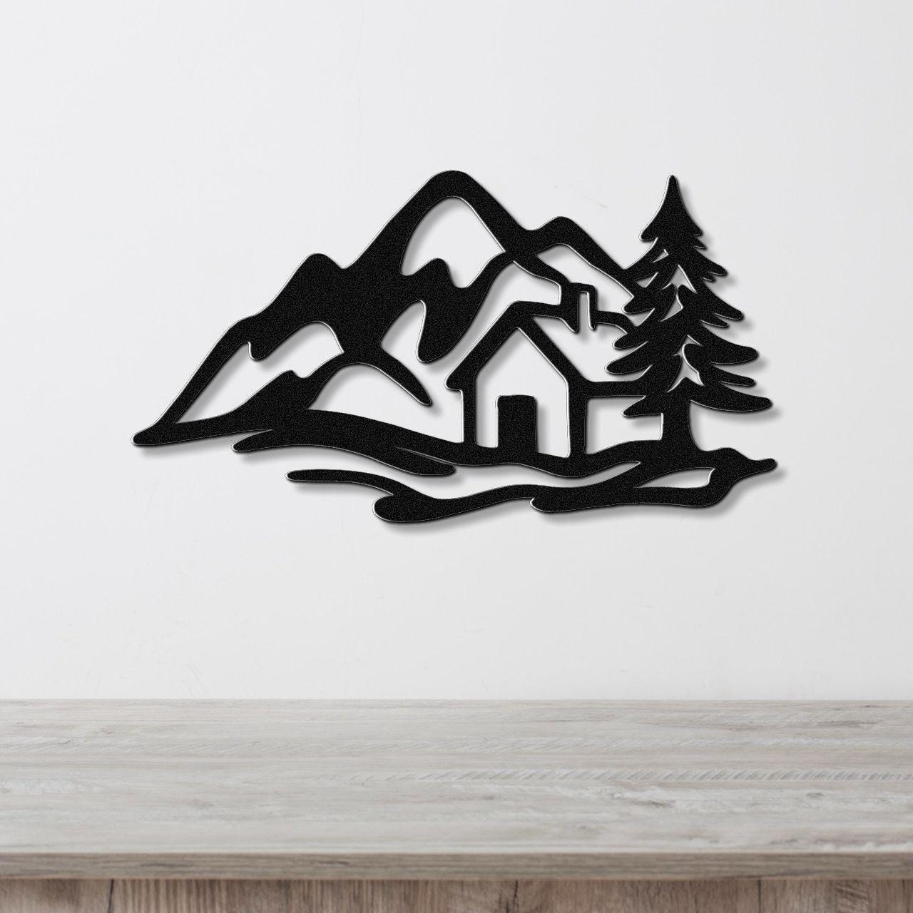Black metal wall art silhouette of a mountain cabin scene with pine trees and peaks, mounted on a white wall above a wooden surface.