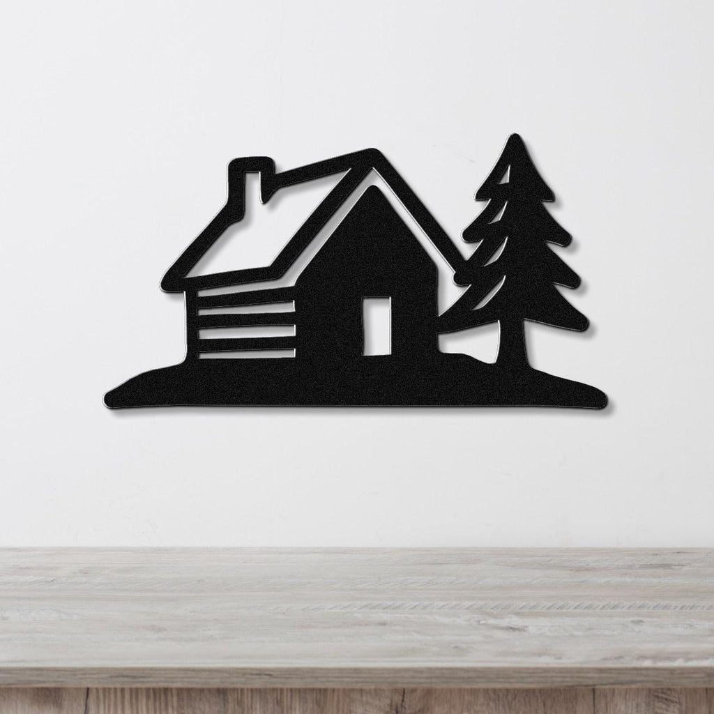 Black metal wall art featuring a cabin and pine tree silhouette, mounted on a white wall above a wooden surface.