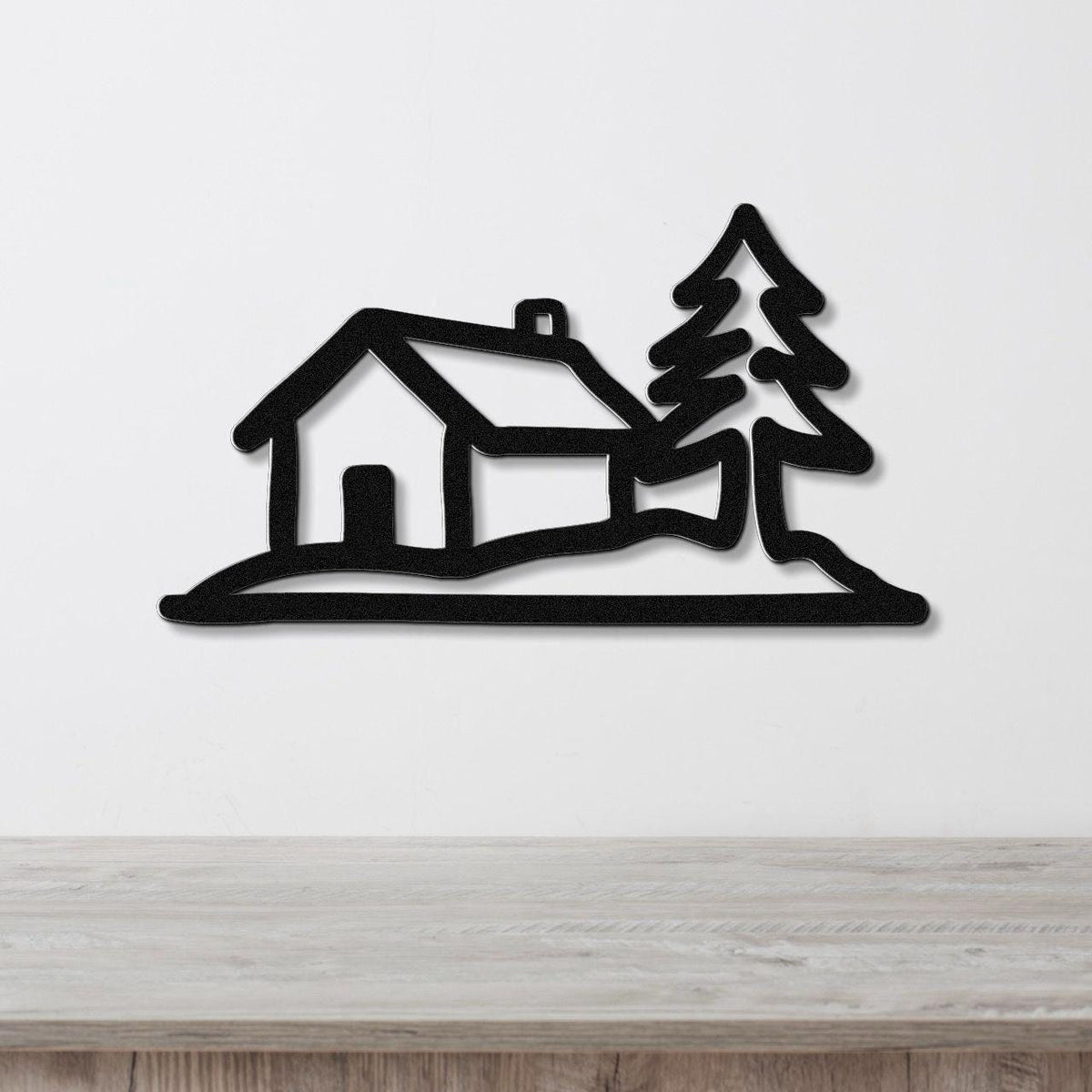 Black metal wall art cutout of a cabin and pine tree silhouette, mounted on a white wall above a light wood counter.