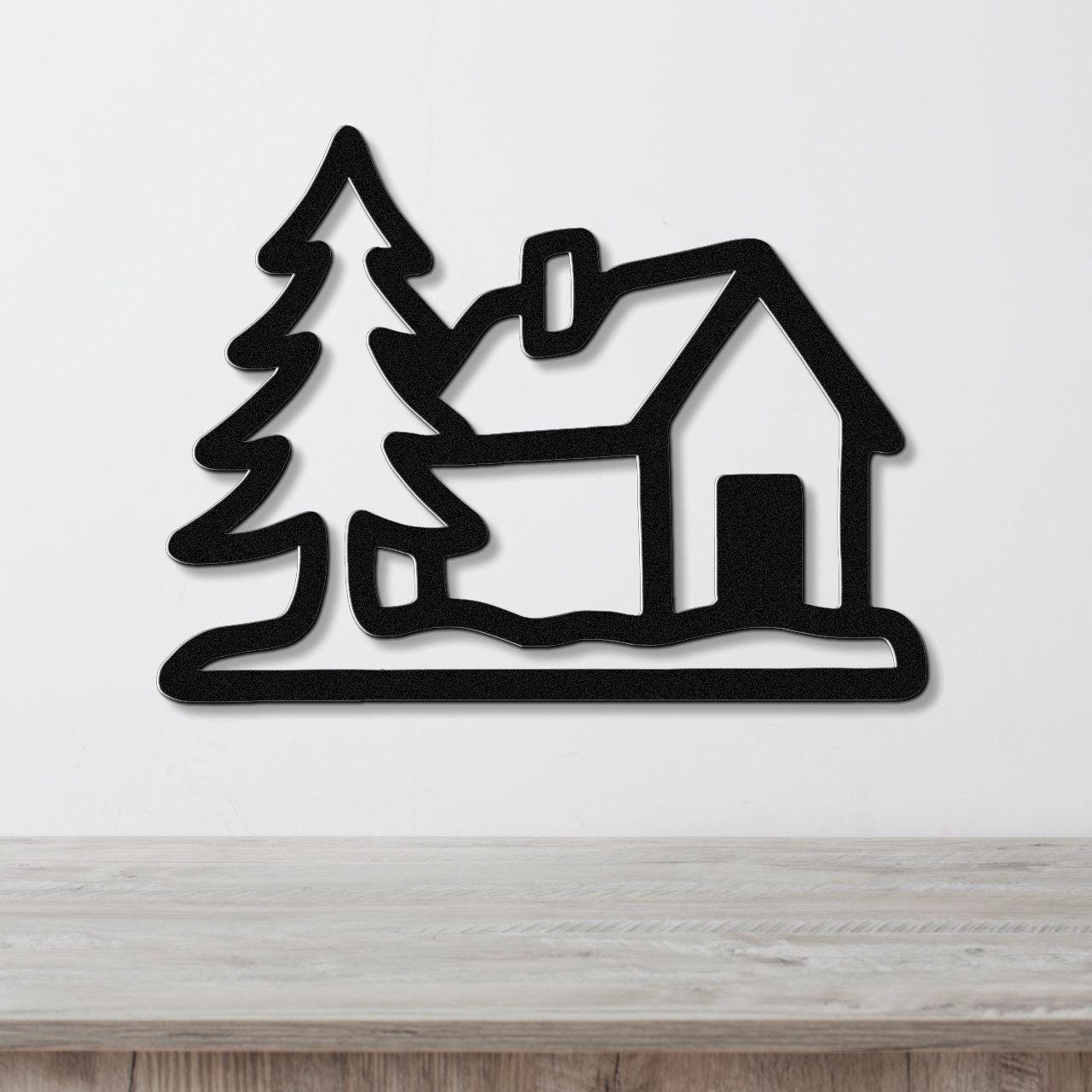 Black metal wall art silhouette of a cabin and pine tree, mounted on a white wall above a wooden surface.