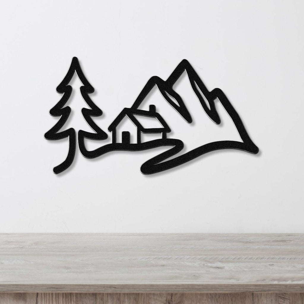 Black metal wall art silhouette of a mountain cabin scene with a pine tree and peaks, mounted on a white wall above a wooden surface.