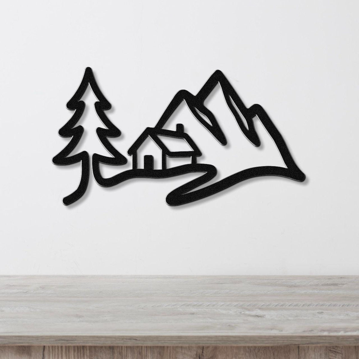 Black metal wall art silhouette of a mountain cabin scene with a pine tree and peaks, mounted on a white wall above a wooden surface.