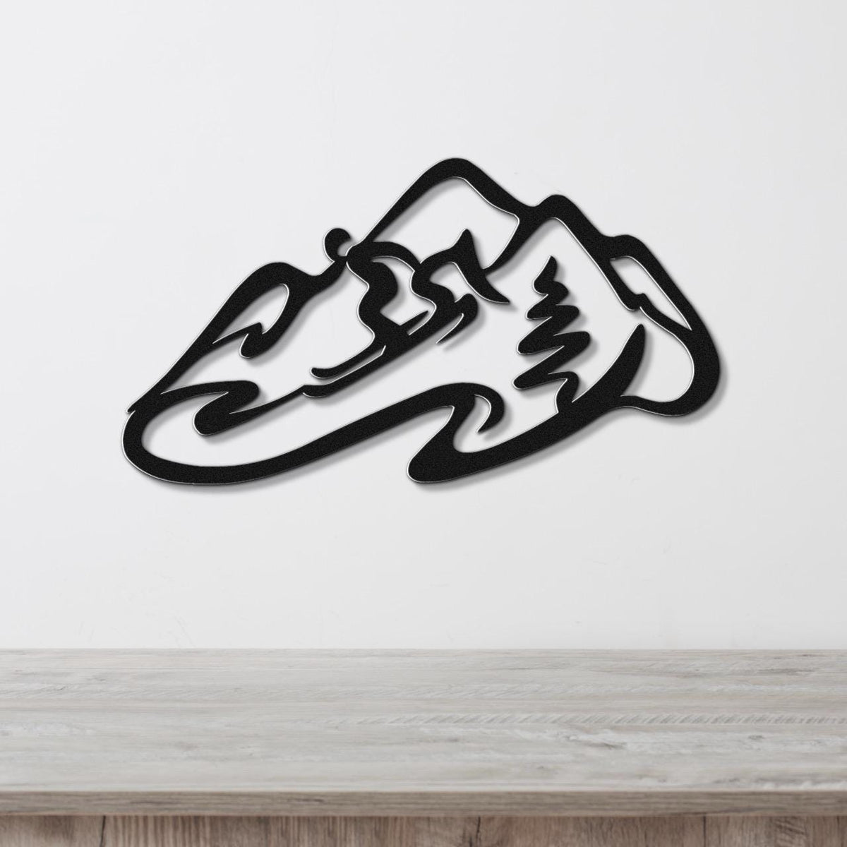 Black metal wall art featuring a minimalist skier silhouette and mountain range cutout, mounted on a white wall above a wooden surface.