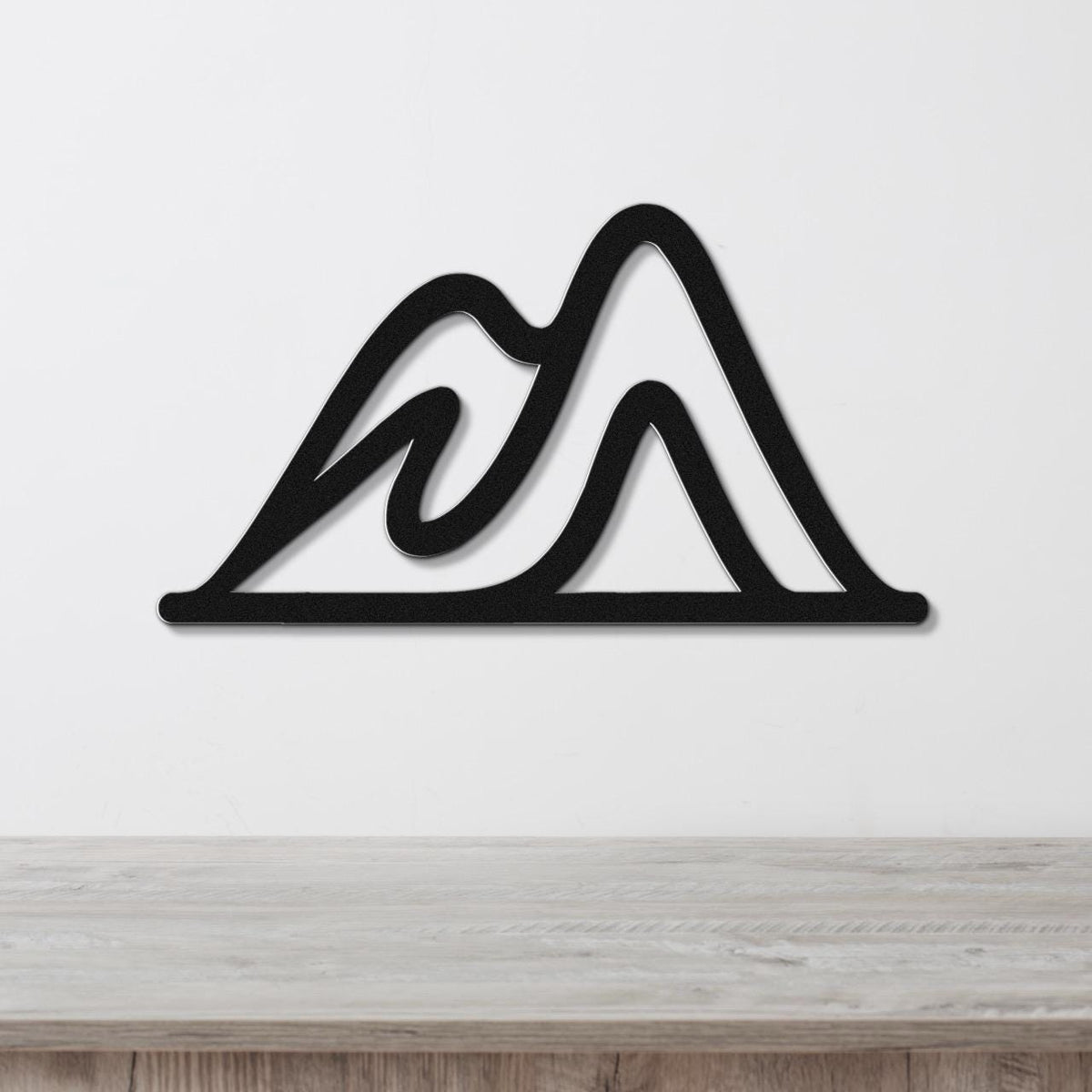 Black metal wall art with a mountain silhouette design, featuring layered peaks and internal cutouts, mounted on a white wall above a wooden surface.