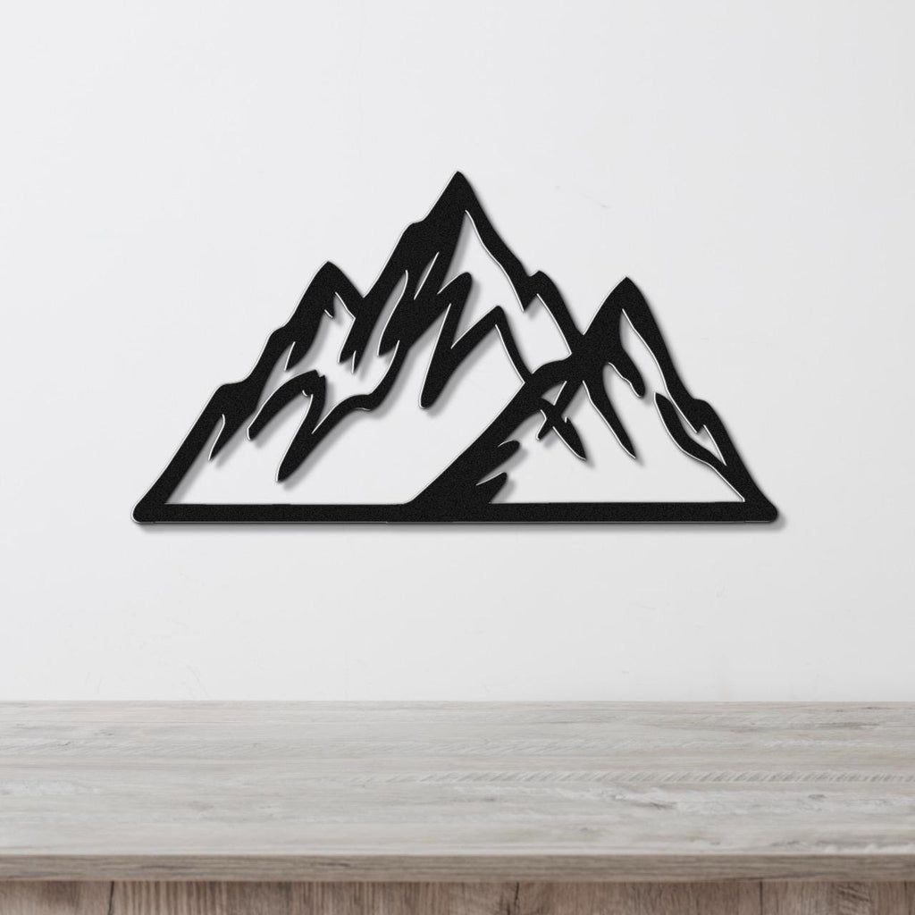 Black metal mountain wall art with layered peak cutouts, mounted on a white wall above a light wood surface.