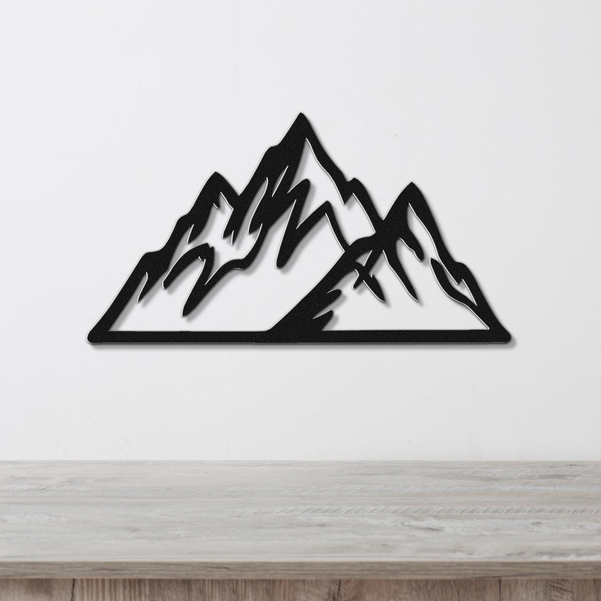 Black metal mountain wall art with layered peak cutouts, mounted on a white wall above a light wood surface.