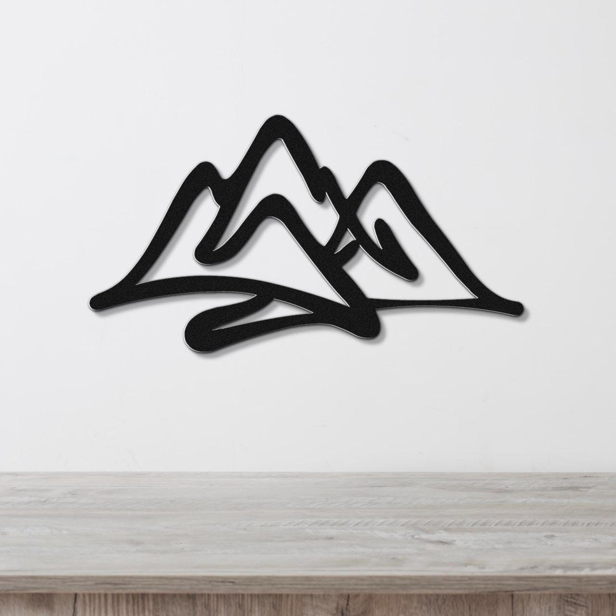 Black metal wall art with a layered mountain silhouette design, mounted on a white wall above a light wood surface.