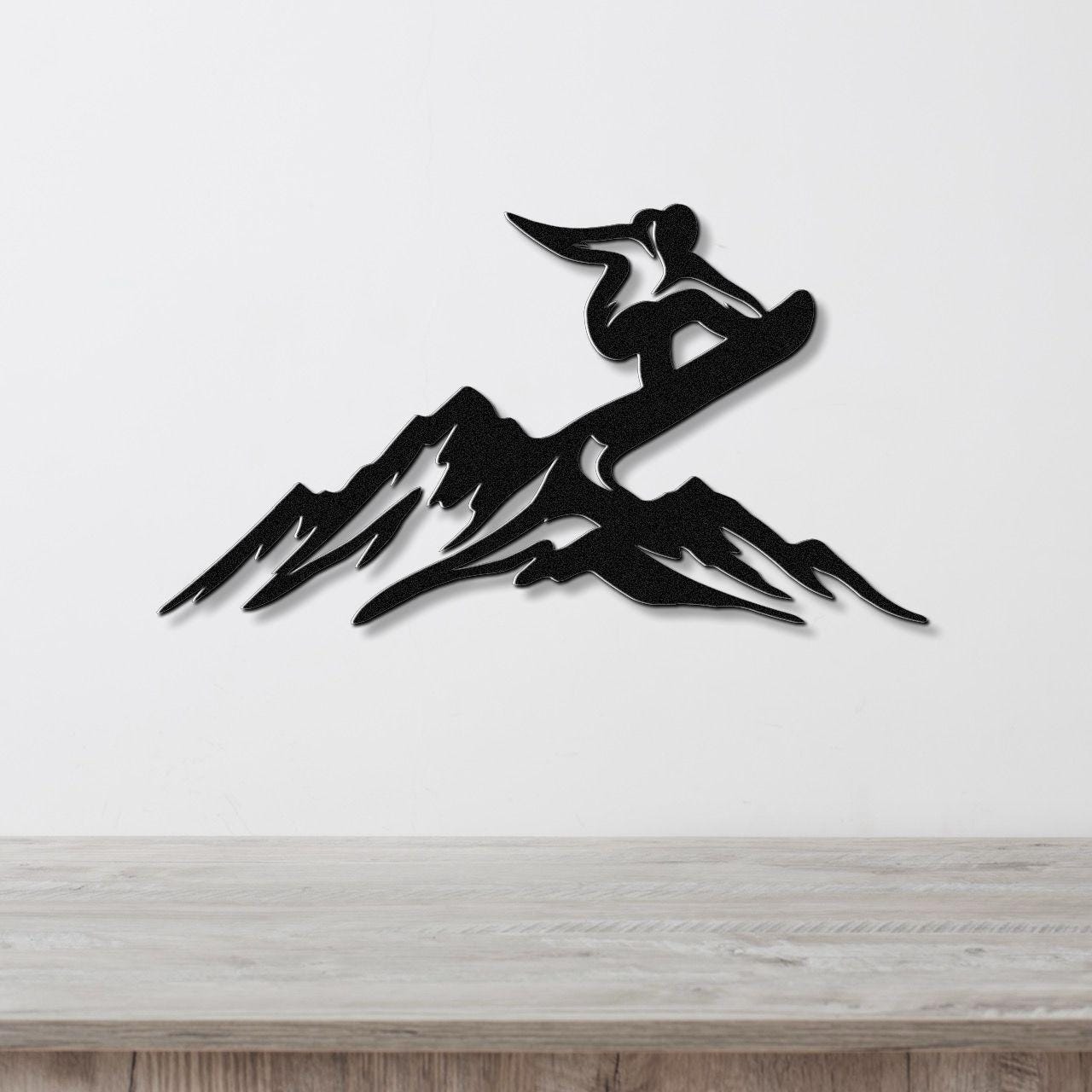 Black metal wall art silhouette of a snowboarder mid-air over mountain peaks, mounted on a white wall above a light wood counter.
