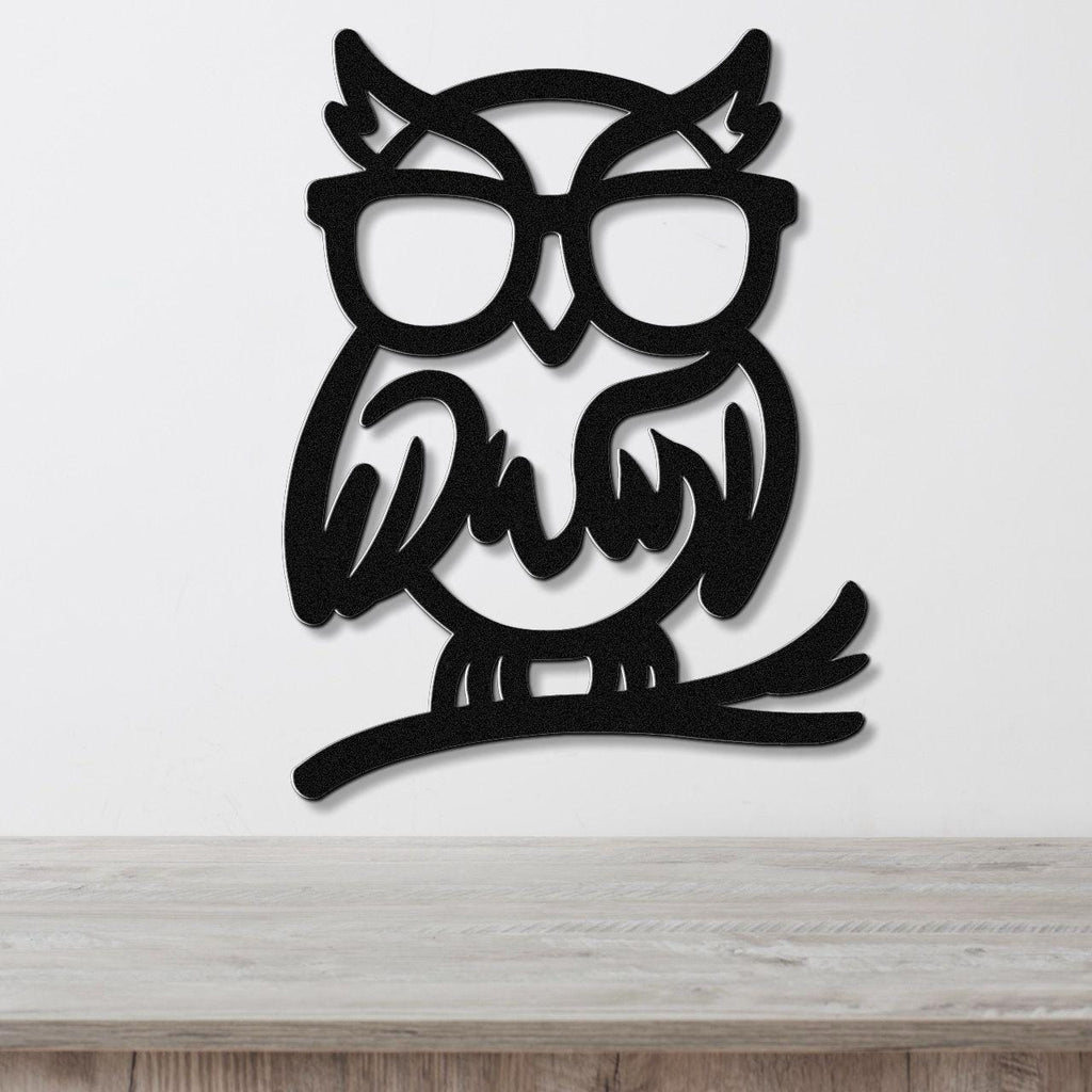 Black metal wall art of a bespectacled owl silhouette with internal cutout details, mounted on a white wall above a light wood surface.