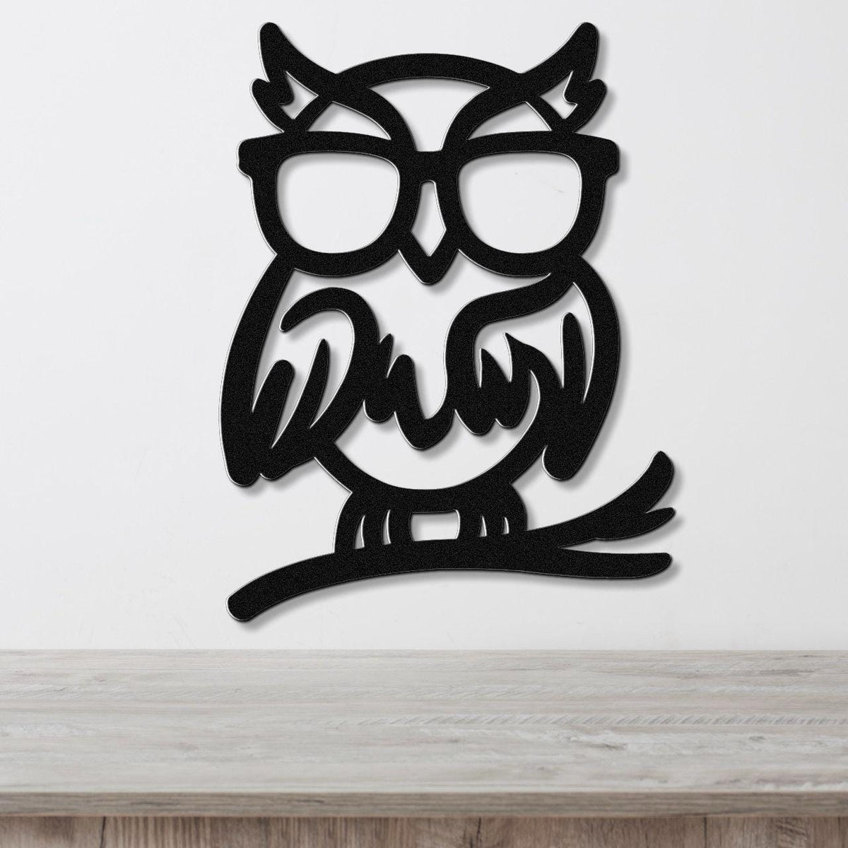 Black metal wall art of a bespectacled owl silhouette with internal cutout details, mounted on a white wall above a light wood surface.